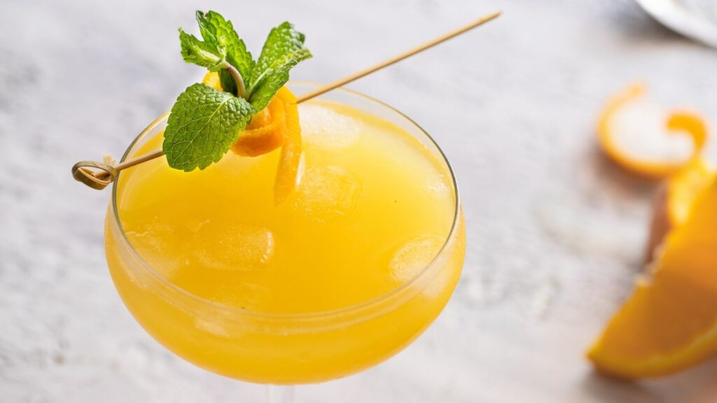 A yellow cocktail with ice is garnished with a sprig of mint and an orange twist on a skewer, served in a coupe glass.