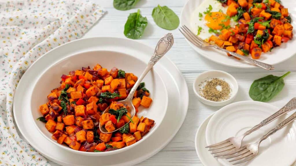 A bowl of sweet potato hash with spinach and red onions, served with a fork, next to plates, forks, spinach leaves, and a small bowl of pepper on a light wooden table.