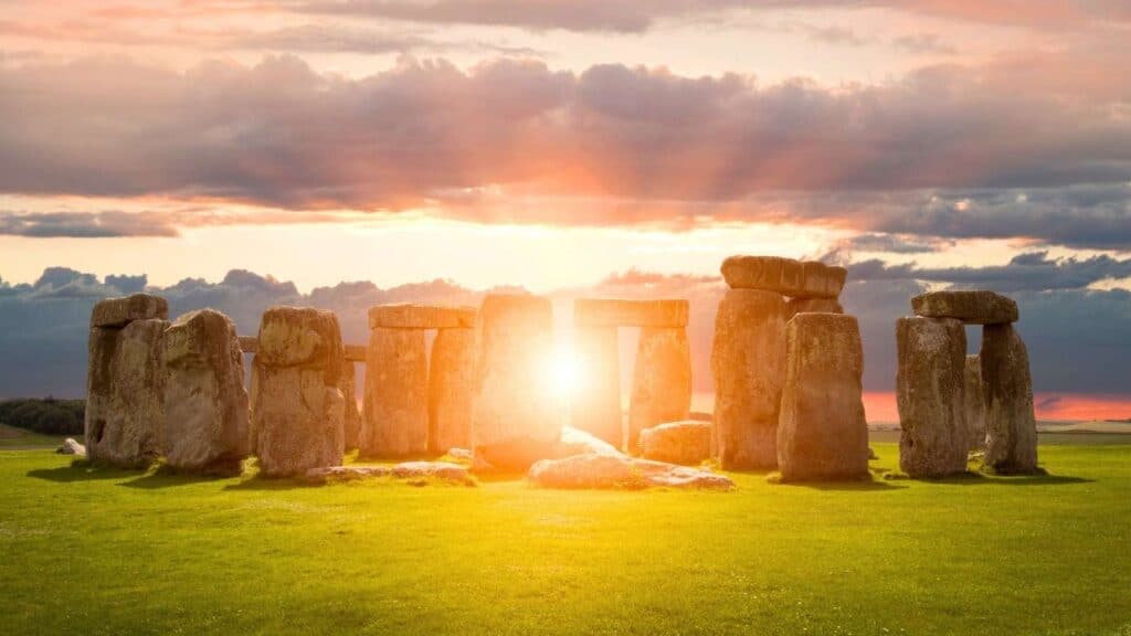 Stonehenge at sunset during the summer solstice, with sunlight shining through the ancient stone monument and a cloudy sky in the background.