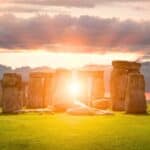 Stonehenge at sunset during the summer solstice, with sunlight shining through the ancient stone monument and a cloudy sky in the background.