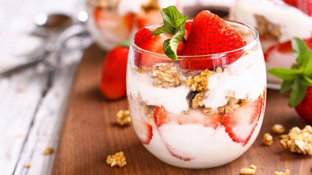 A glass filled with layers of yogurt, granola, and sliced strawberries, garnished with a sprig of mint, sits on a wooden surface.