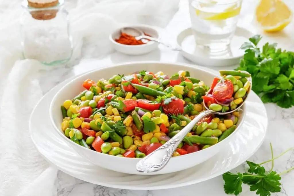 A white bowl of colorful vegetable salad with corn, green beans, edamame, and tomatoes, served with a spoon on a plate. A glass of water and seasonings are in the background.