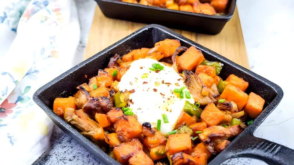 A black square dish filled with roasted sweet potatoes, vegetables, and shredded meat, topped with a poached egg and garnished with chopped green onions.