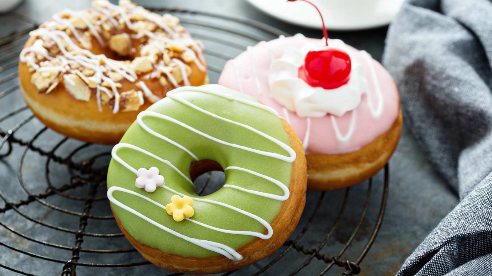 Three decorated donuts on a wire rack: one with nuts and icing, one with green icing and flower decorations, and one with pink icing, whipped cream, and a cherry—perfect treats to celebrate National Donut Day.
