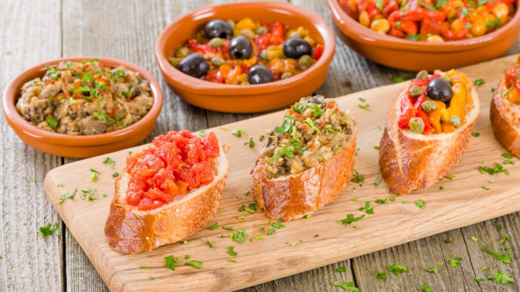 A wooden board with assorted bruschetta topped with tomatoes, eggplant, and peppers, with small bowls of mixed vegetable appetizers in the background.