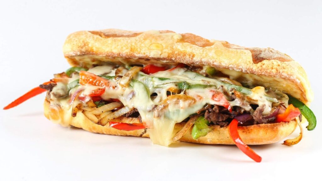 A toasted sandwich roll filled with sliced beef, melted cheese, onions, and red and green bell peppers, inspired by the classic Philadelphia style, set against a white background.