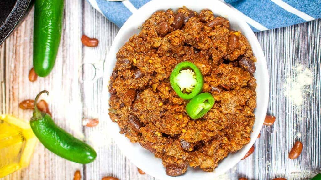 A bowl of chili with ground meat and beans, topped with sliced jalape&ntilde;os, sits on a wooden surface with whole jalape&ntilde;os and scattered beans nearby.