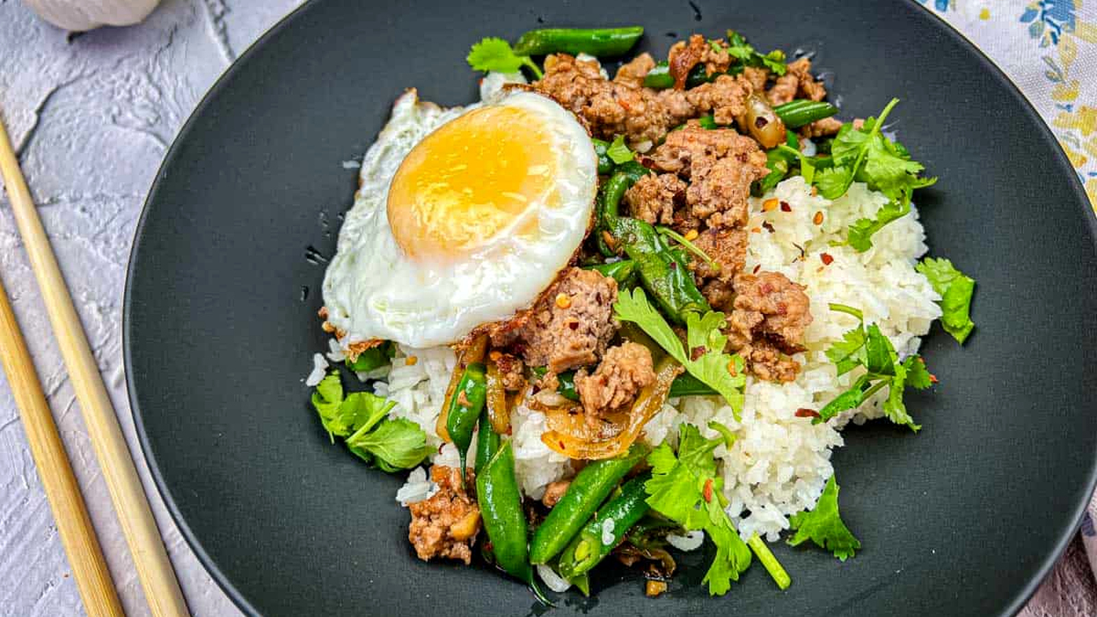 A plate of Thai pork & Green beans with rice, topped with a fried egg, served with chopsticks on the side.