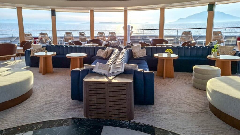 Lounge area with blue sofas, round tables, and books, overlooking a sunlit deck and ocean through large windows—perfect for relaxing during your Windstar Alaska cruise.