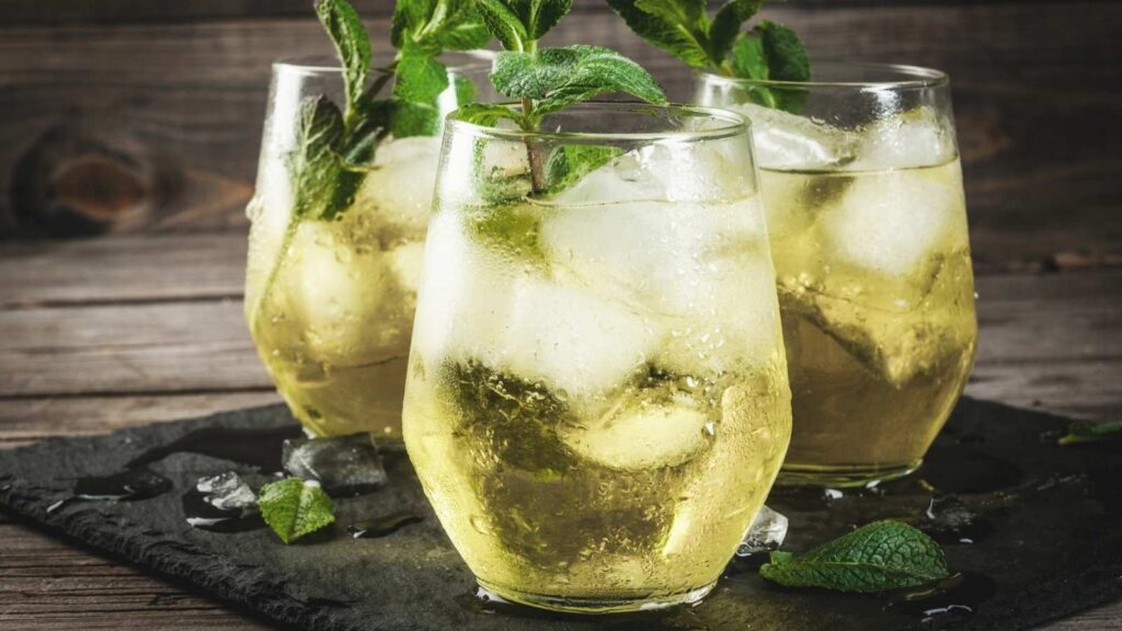 Three glasses of iced greenish-yellow drink garnished with fresh mint leaves sit on a slate surface, with scattered ice cubes and mint around them.