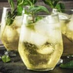 Three glasses of iced greenish-yellow drink garnished with fresh mint leaves sit on a slate surface, with scattered ice cubes and mint around them.
