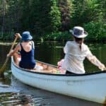 Two people in hats paddle a canoe on a calm, tree-lined lake on a sunny day.