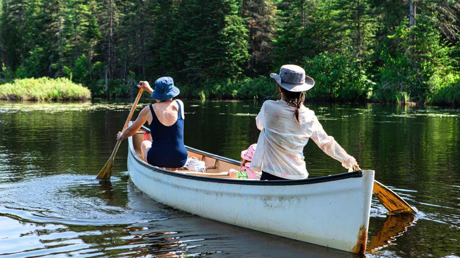 Two people in hats paddle a canoe on a calm, tree-lined lake on a sunny day.