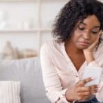 A woman sits on a couch, looking at her smartphone with her head resting on her hand, appearing thoughtful or concerned—perhaps reflecting on the effects of the friendship recession.