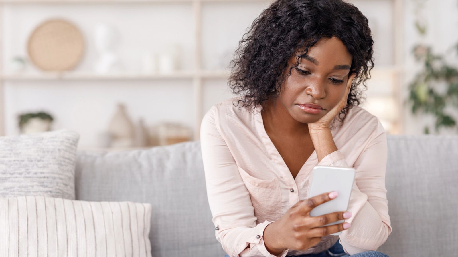 A woman sits on a couch, looking at her smartphone with her head resting on her hand, appearing thoughtful or concerned—perhaps reflecting on the effects of the friendship recession.