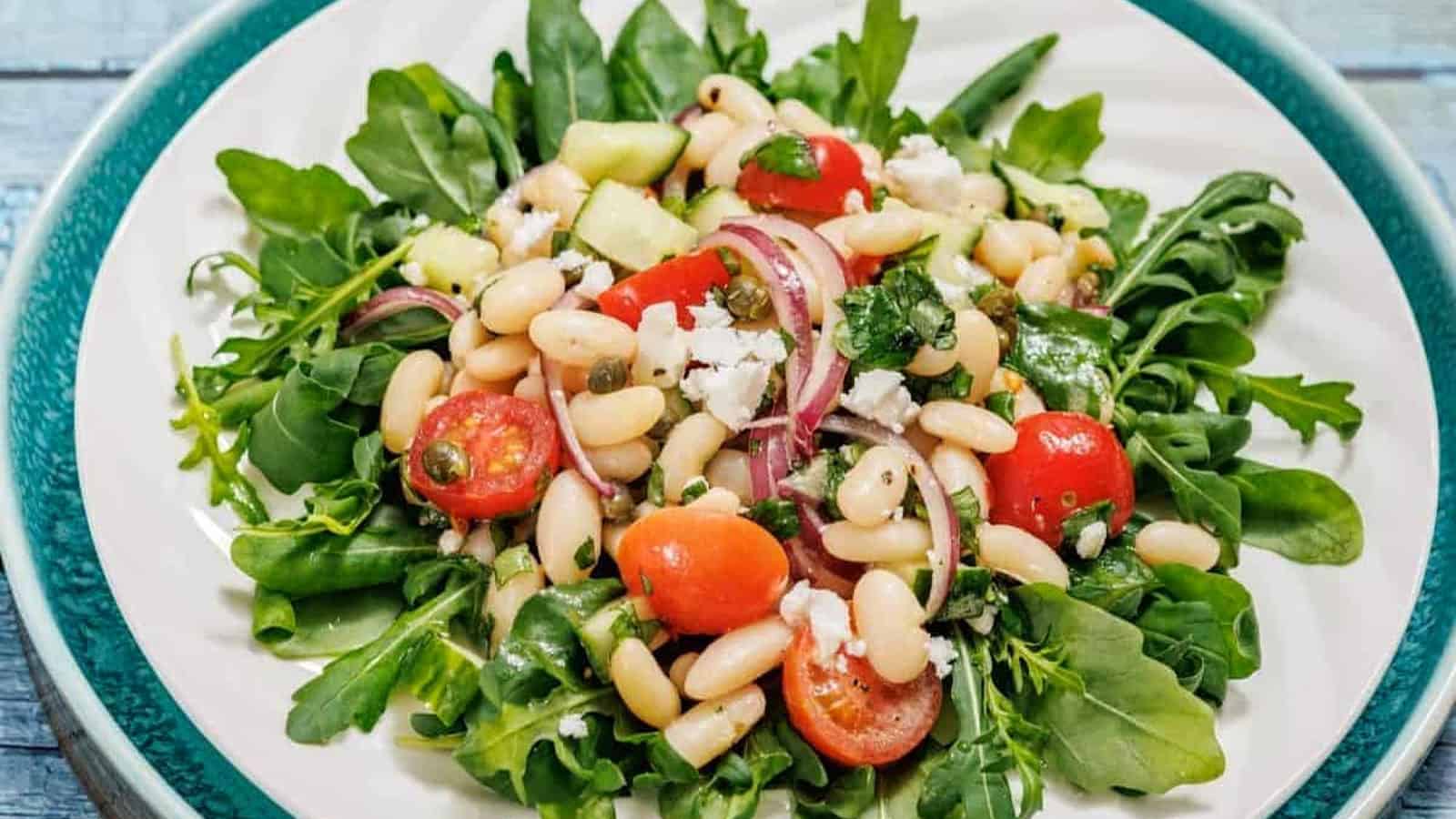 A plate of White Bean Salad with arugula, white beans, cherry tomatoes, red onion, cucumber, and crumbled cheese.
