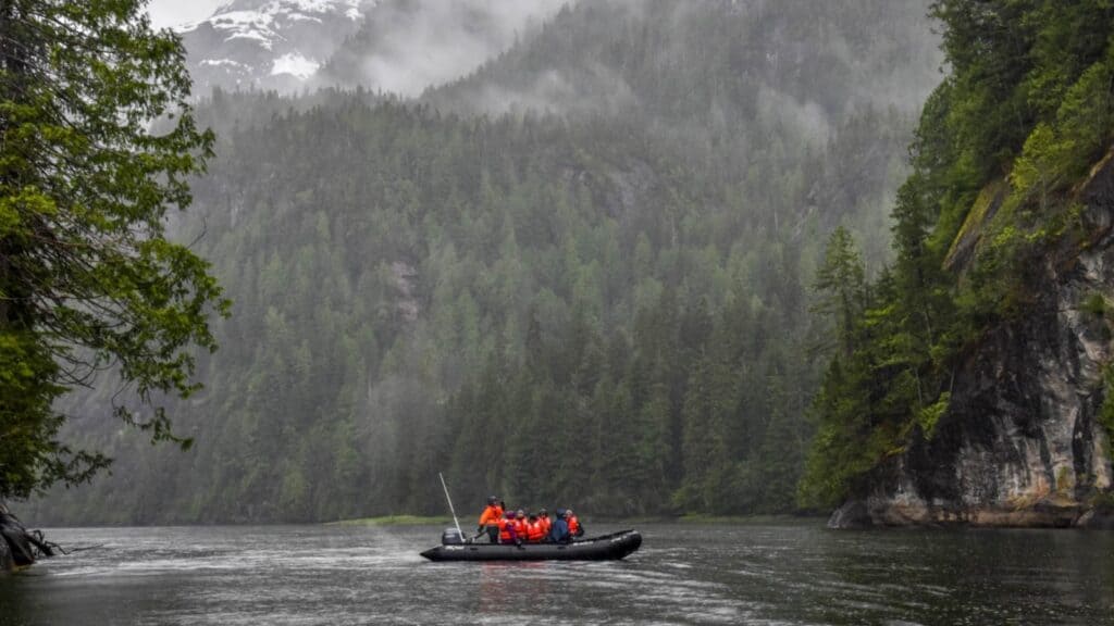 A group of people in orange jackets ride an inflatable boat on a river, surrounded by dense, misty forest and mountains—an adventure reminiscent of a Windstar Alaska cruise.