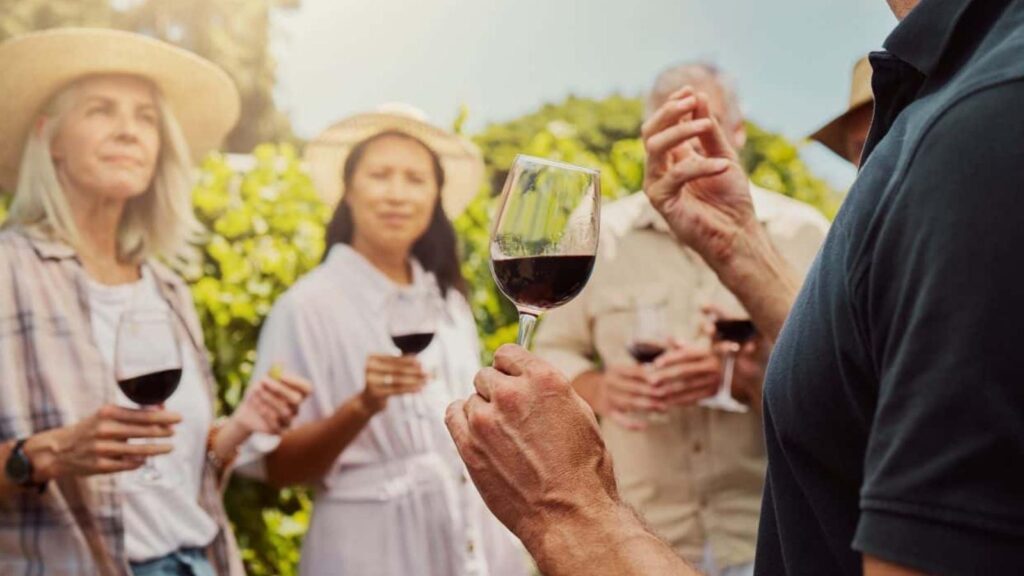 A group of people standing outdoors holding glasses of red wine during one of their wine trips, listening to a person speaking, with greenery in the background.