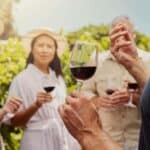 A group of people standing outdoors holding glasses of red wine during one of their wine trips, listening to a person speaking, with greenery in the background.