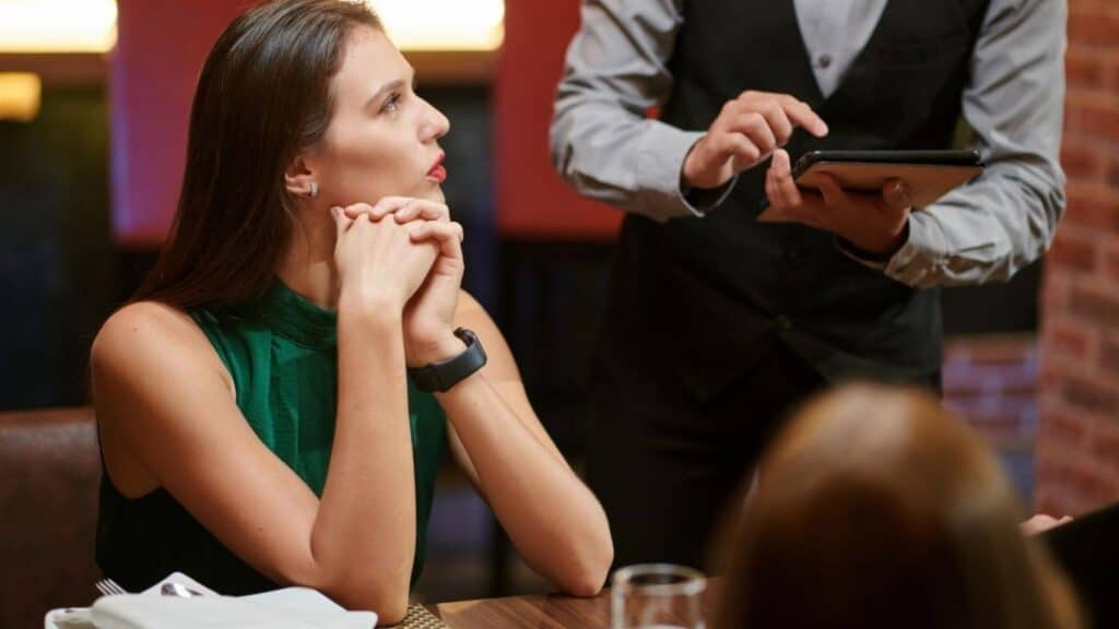 A woman sits at a restaurant table with her hands clasped, looking to the side as a server holding a tablet takes an order.