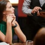 A woman sits at a restaurant table with her hands clasped, looking to the side as a server holding a tablet takes an order.