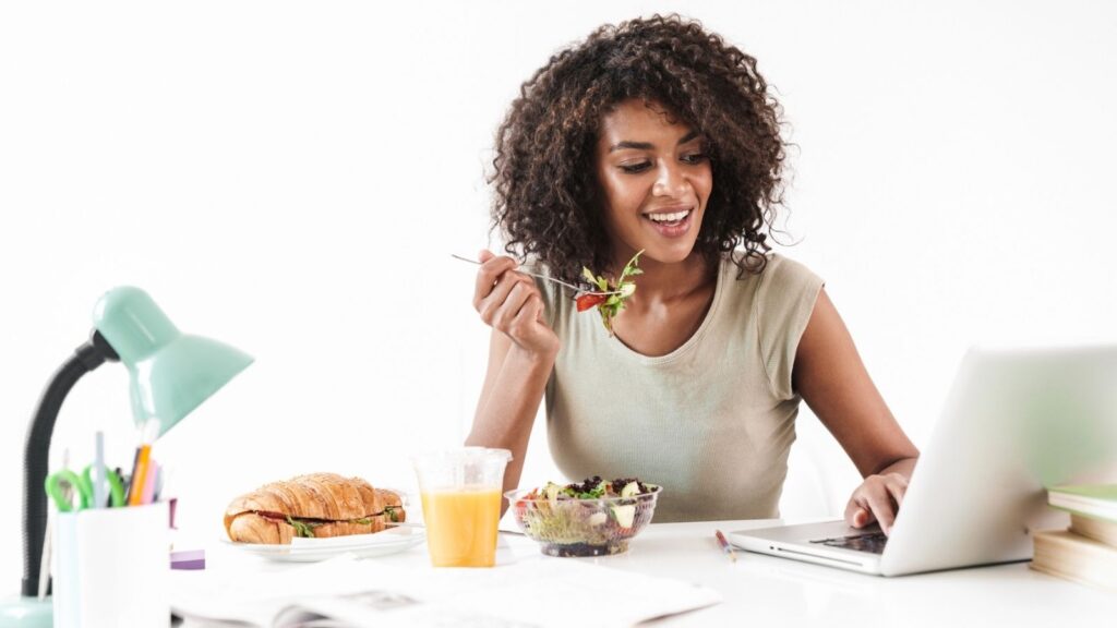 On national work from home day, a woman sits at her desk enjoying a salad while working on her laptop, with a croissant sandwich, orange juice, and a lamp by her side.