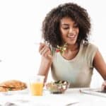 On national work from home day, a woman sits at her desk enjoying a salad while working on her laptop, with a croissant sandwich, orange juice, and a lamp by her side.