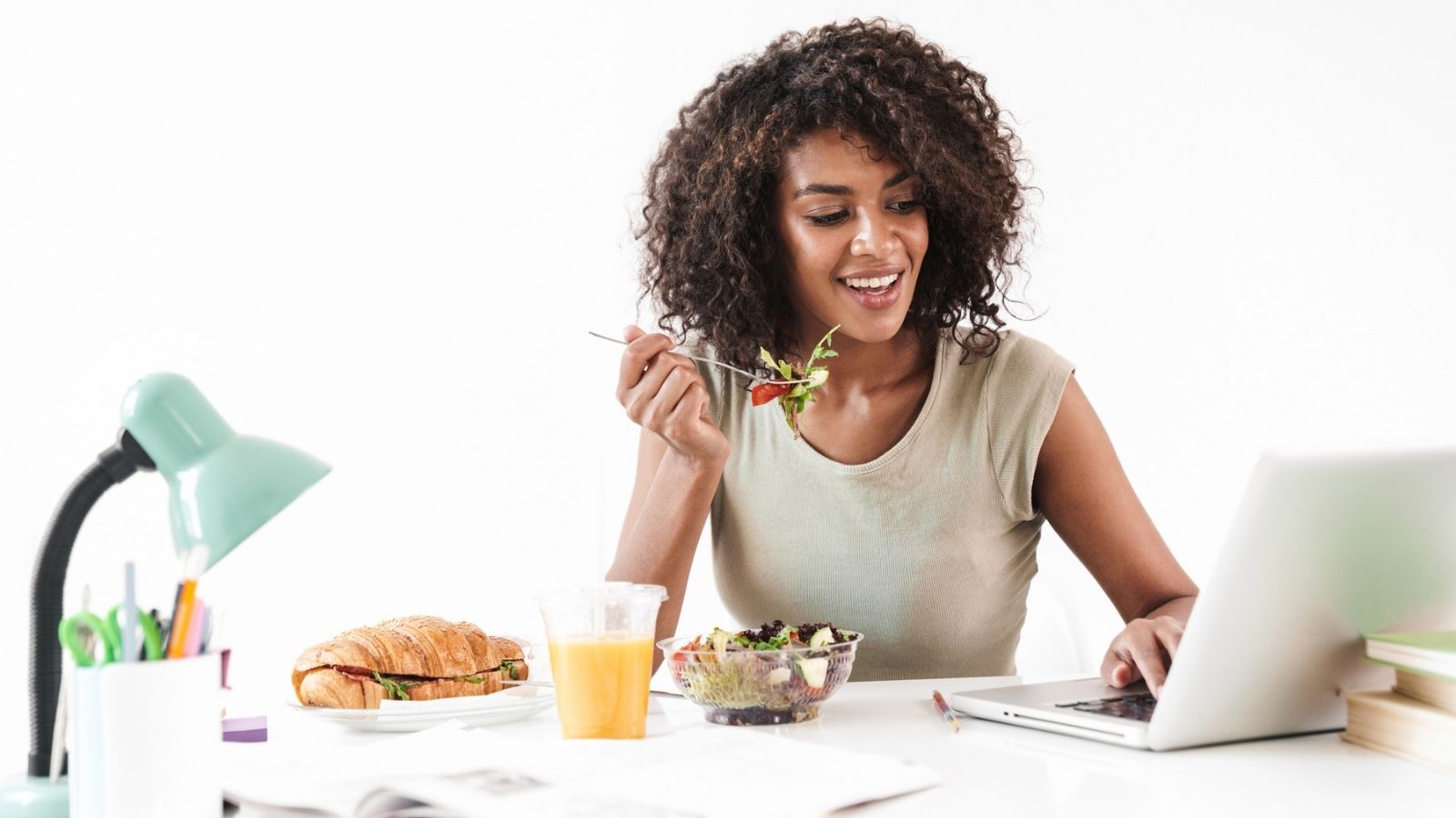 On national work from home day, a woman sits at her desk enjoying a salad while working on her laptop, with a croissant sandwich, orange juice, and a lamp by her side.