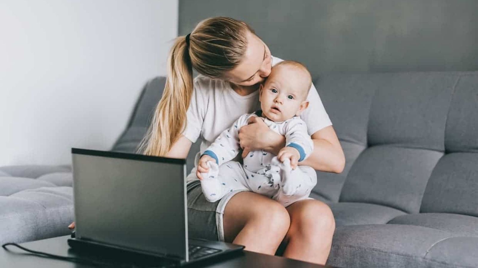 A woman sits on a couch holding a baby in her lap with a laptop on the table in front of them.