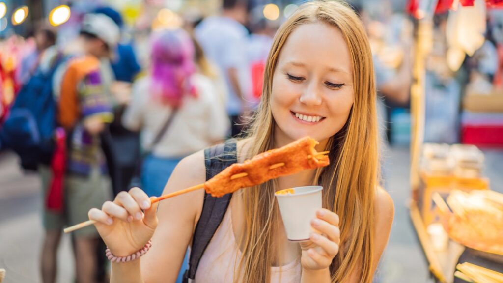 A woman at an outdoor market smiles while snackpacking, holding a cup and enjoying a deep-fried food item on a skewer.