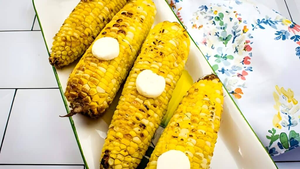 Four grilled corn cobs with pat of butter on each, served on a rectangular white tray with a floral napkin nearby.