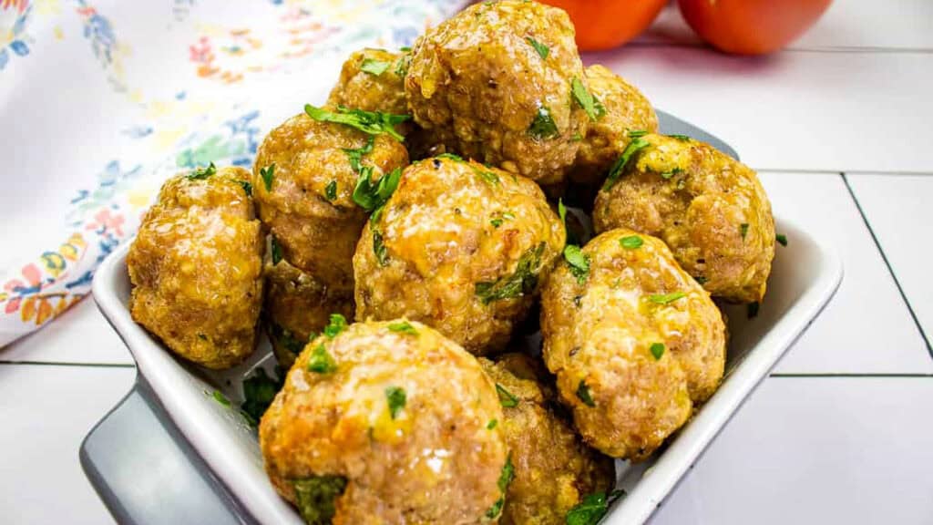 A bowl filled with several cooked meatballs garnished with chopped herbs, set on a white tiled surface with tomatoes and a floral cloth in the background.