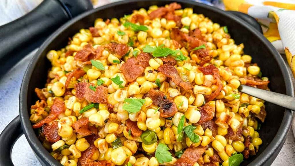 A bowl of fried corn mixed with crispy bacon pieces and garnished with chopped parsley, with a spoon resting in the dish.