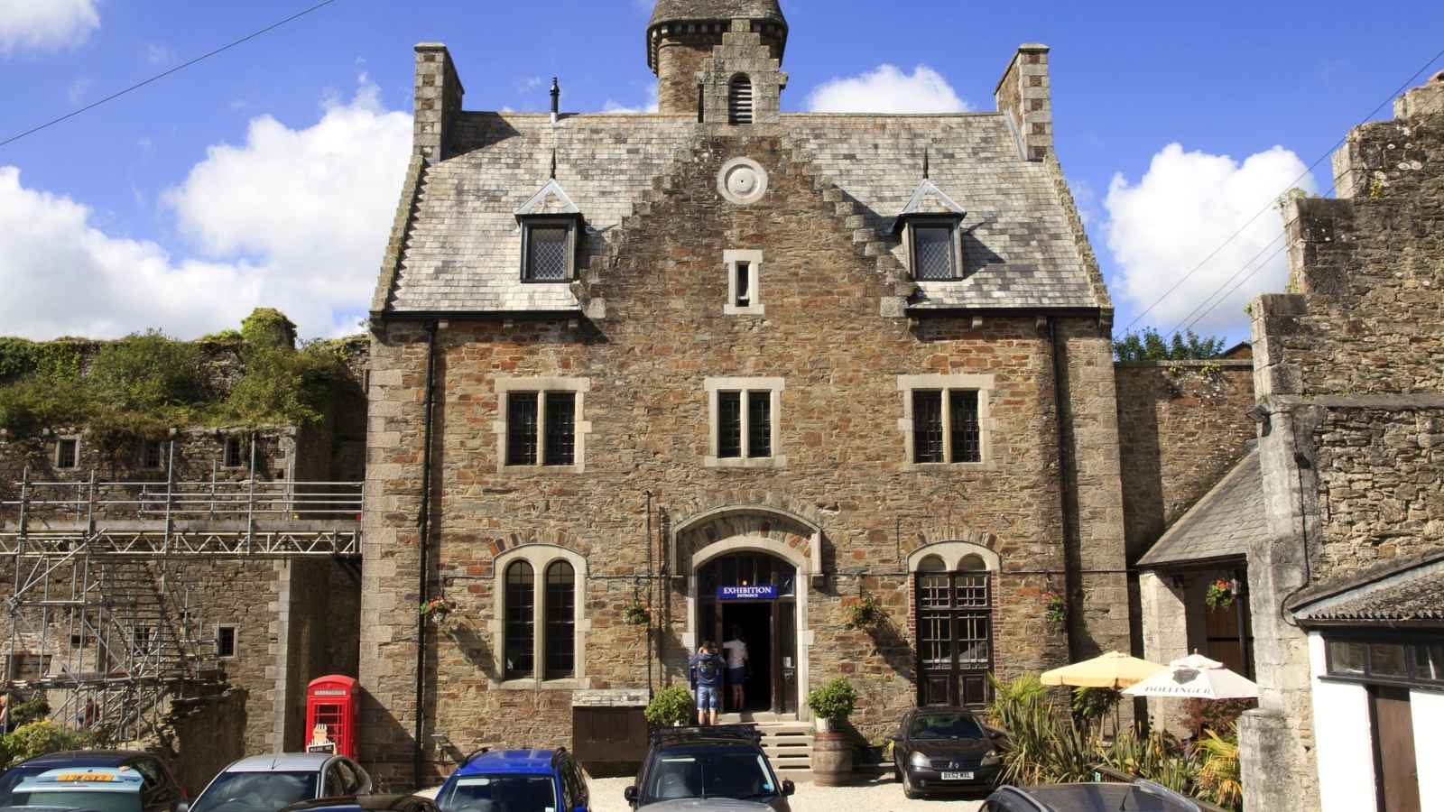 Stone building with a steep slate roof made from salvaged stays, arched windows, and central clock tower; several parked cars and people at entrance; blue sky with clouds.