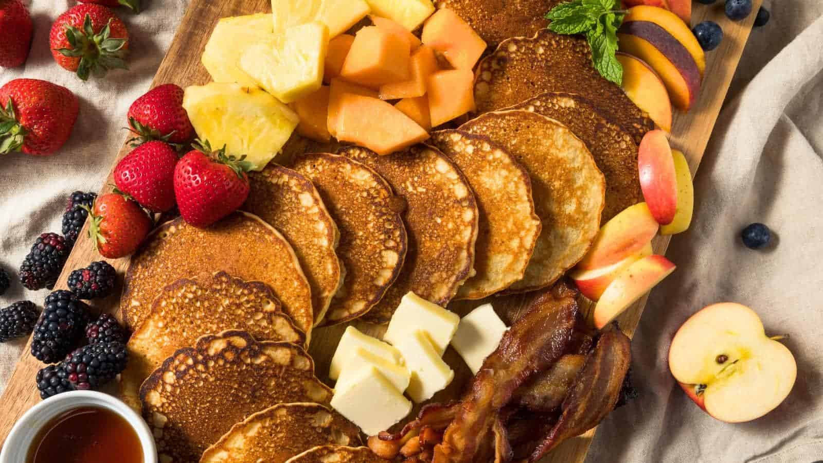 A wooden board with pancakes, butter, bacon, fresh strawberries, blackberries, pineapple, cantaloupe, apple slices, blueberries, and a small bowl of syrup.