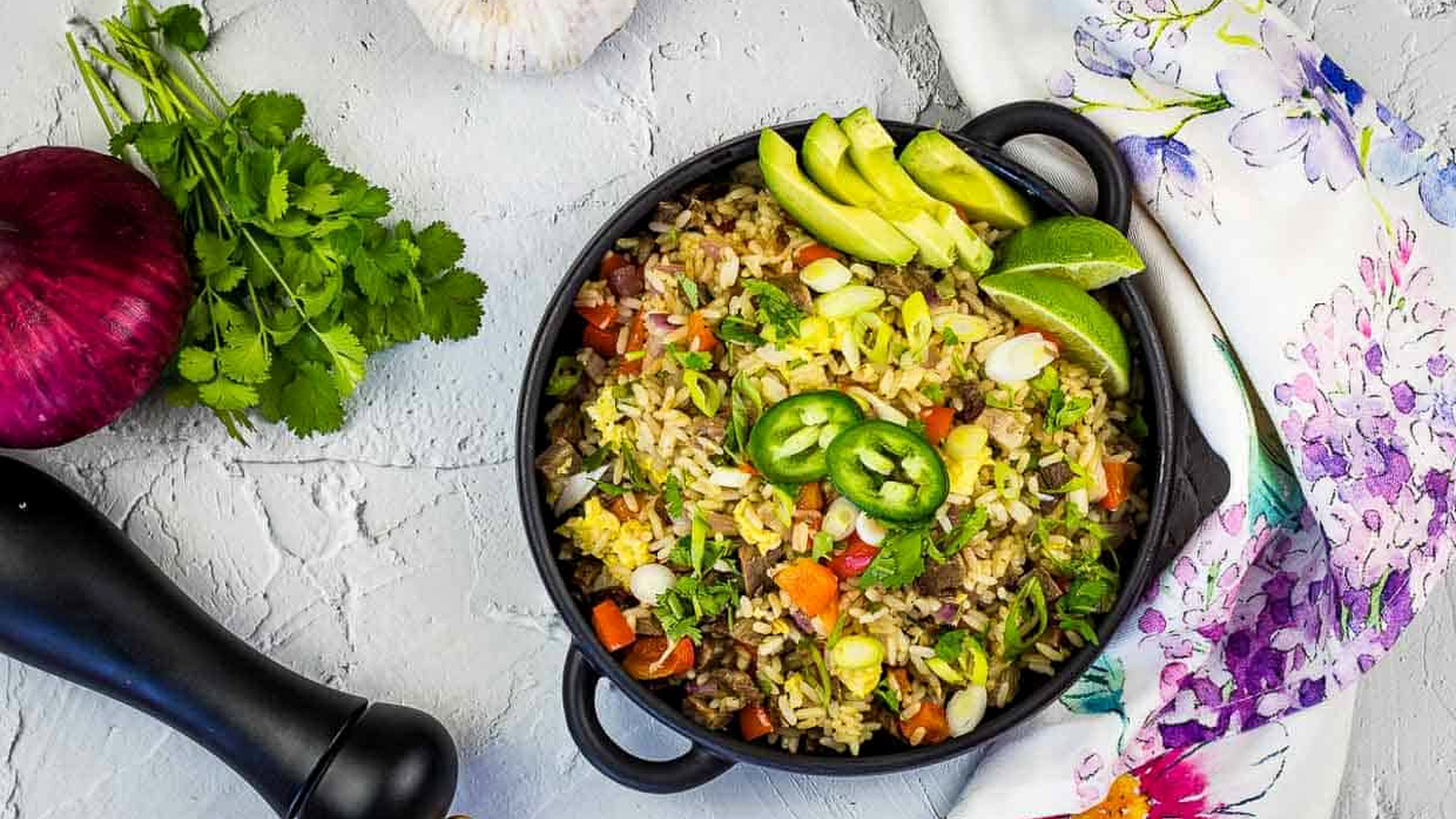 A cast-iron pan filled with Brisket Fried Rice, garnished with sliced avocado, lime wedges, jalape&ntilde;o slices, and fresh cilantro.