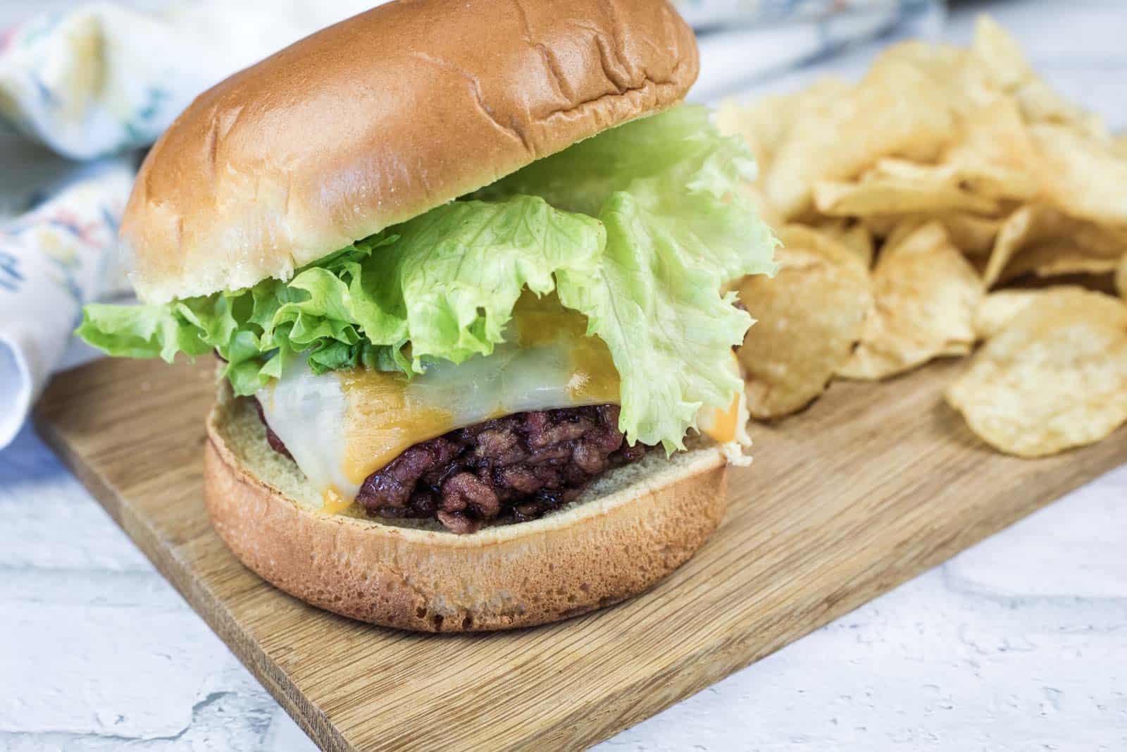 A smoked burger with lettuce and a slice of cheddar cheese on a bun, served with potato chips on a wooden board.