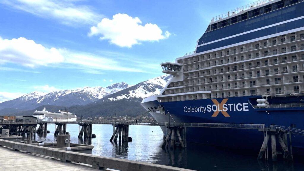 Two large cruise ships docked at a pier, framed by snow-capped mountains and a partly cloudy sky. The ship in the foreground, "Celebrity Solstice," hints at the luxury and refined experiences, like Celebrity Cruises wine tastings onboard.