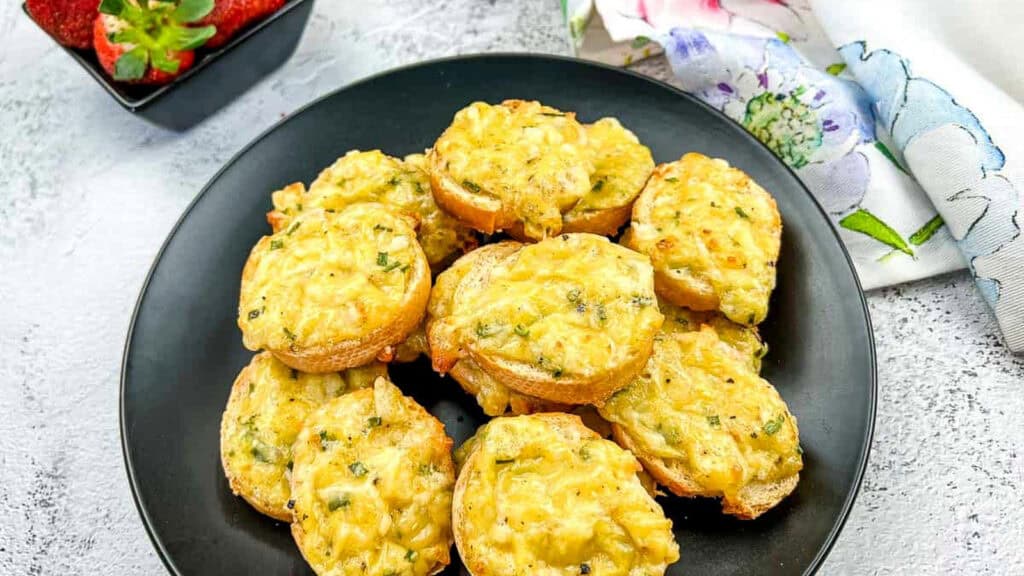 Eight pieces of toasted bread topped with melted cheese and herbs are arranged on a black plate; strawberries and a floral napkin are nearby.
