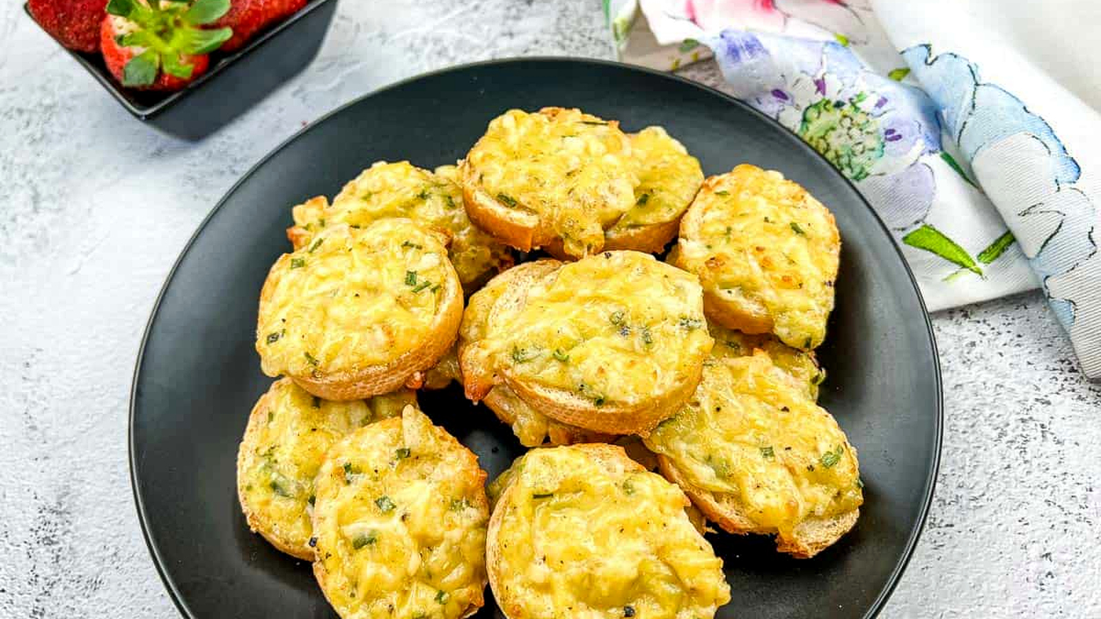 Bite-sized Cheese Toasties on a black plate with a small bowl of strawberries and a floral napkin nearby.