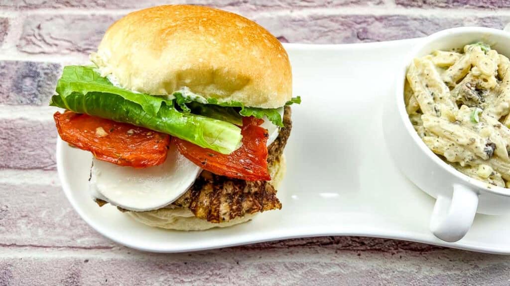A grilled chicken sandwich with lettuce, tomato, and cheese on a bun is served next to a cup of creamy pasta on a white plate.