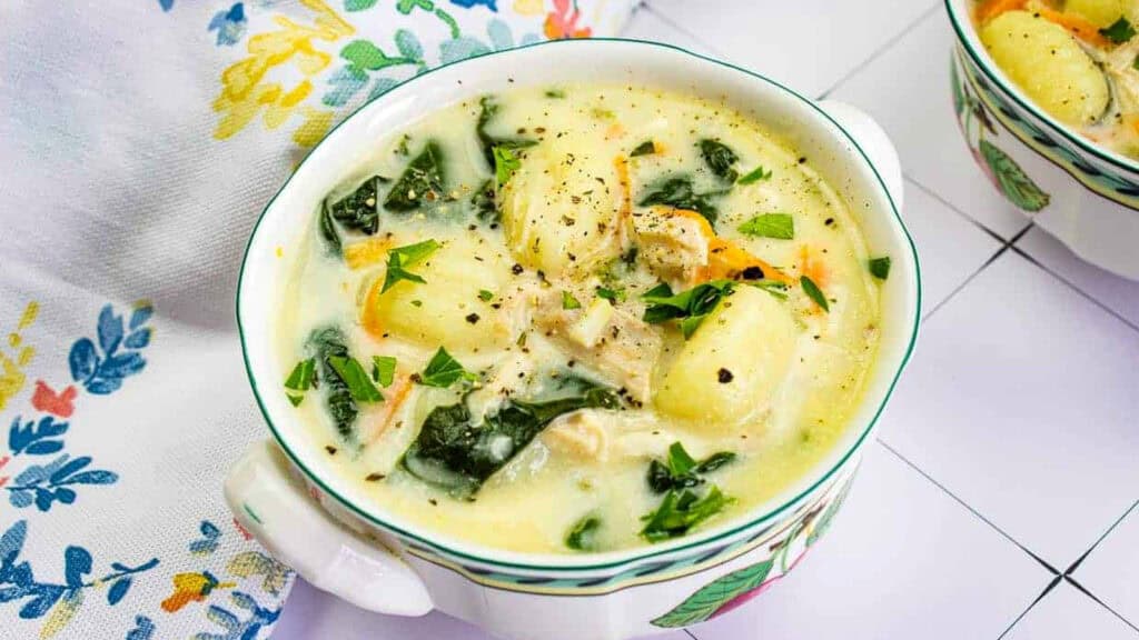A bowl of creamy chicken gnocchi soup with spinach, carrots, and black pepper, garnished with chopped parsley.