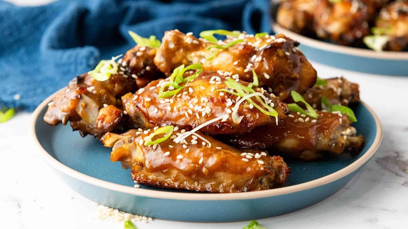 A plate of glazed chicken wings garnished with sesame seeds and sliced green onions, served on a blue dish.