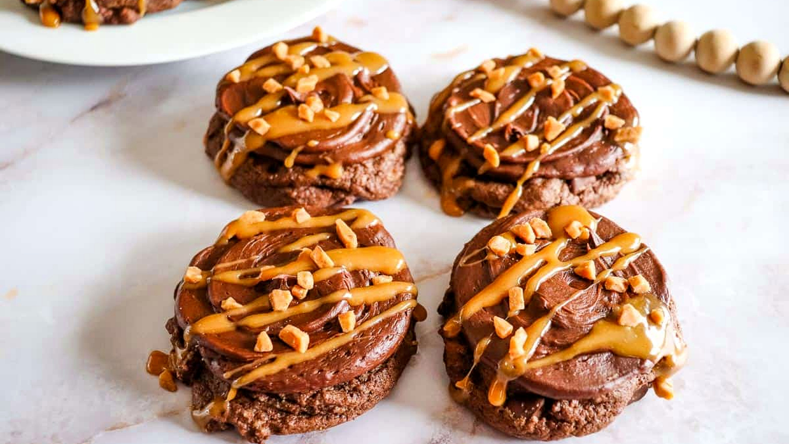 Chocolate Caramel Cookies with frosting, caramel drizzle, and chopped nuts on a white marble surface.