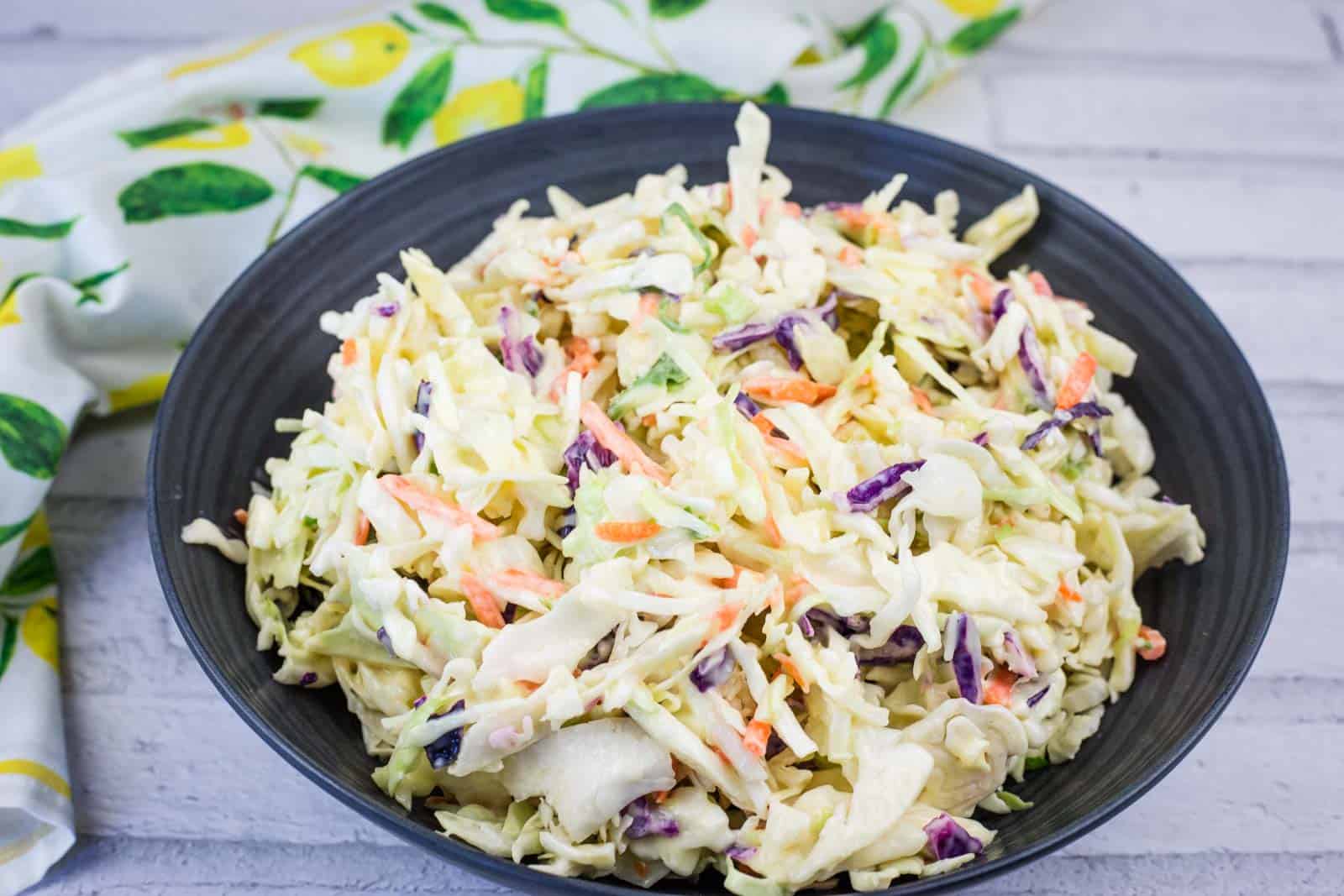 A black bowl filled with creamy coleslaw made of shredded cabbage, carrots, and purple cabbage, placed on a light-colored surface with a patterned cloth nearby.