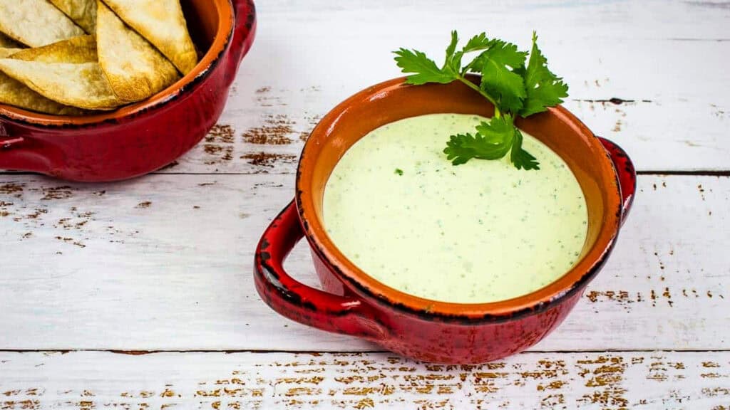 A red ceramic bowl filled with green creamy dip garnished with cilantro, next to a bowl of triangular potato wedges on a white wooden surface.
