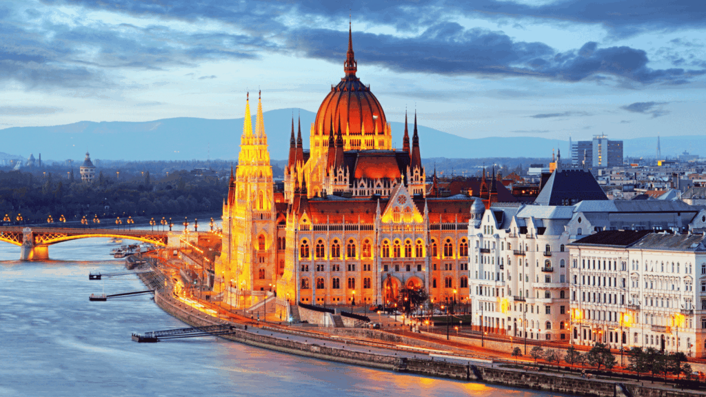 The illuminated Hungarian Parliament Building sits by the Danube River in Budapest at dusk, with surrounding historical architecture and a bridge in the background.