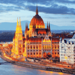 The illuminated Hungarian Parliament Building sits by the Danube River in Budapest at dusk, with surrounding historical architecture and a bridge in the background.