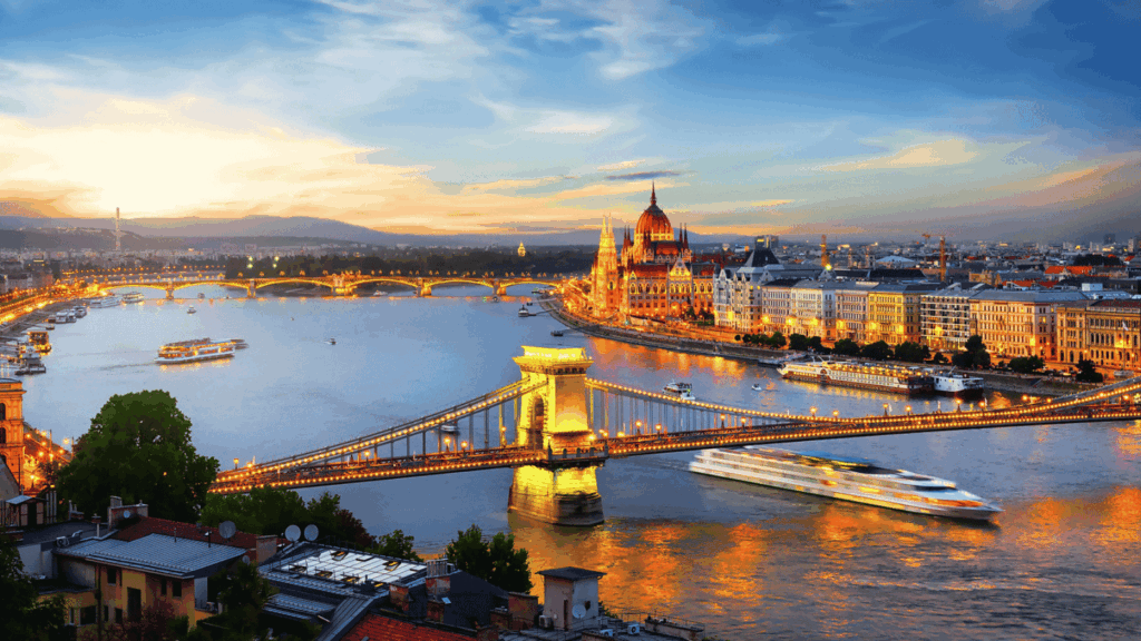 A view of the Chain Bridge over the Danube River in Budapest at sunset, with the Hungarian Parliament Building and city lights in the background.