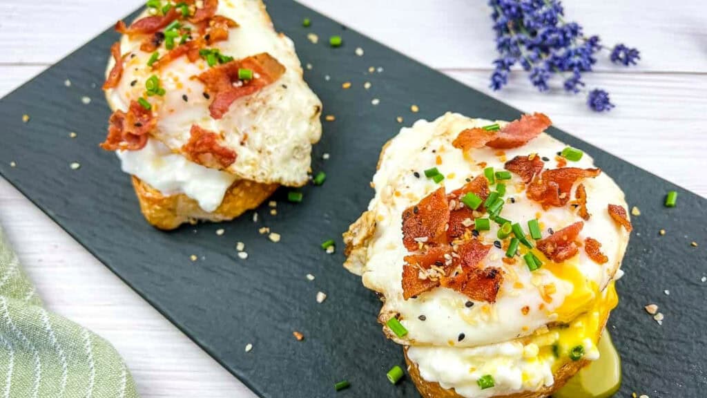 Two pieces of toast topped with fried eggs, crumbled bacon, chopped chives, and seasoning are served on a black rectangular plate.